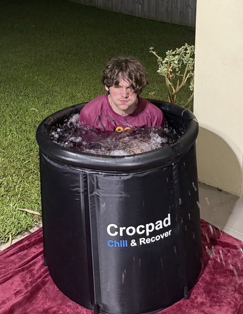A young footballer in a maroon jersey sitting shoulder-deep in a black Crocpad Chill & Recover portable ice bath on backyard turf at night